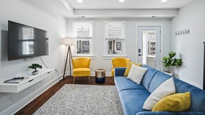 Ein modernes Wohnzimmer mit Fernseher, gelben Sesseln, einem blauen Sofa und einer kleinen Pflanze.