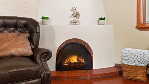 A fireplace with a brick surround, a leather sofa with a throw pillow, and two small potted plants on a shelf above the fireplace.