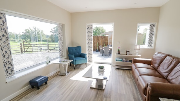 A spacious living room with a brown leather sofa, a blue armchair, and a white coffee table. Large windows offer a view of a fenced outdoor area. - Southend (Malvern)