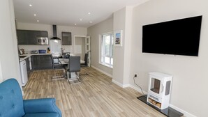A modern living room with a flat-screen TV, a blue armchair, and a kitchen area with stainless steel appliances. - Southend (Malvern)