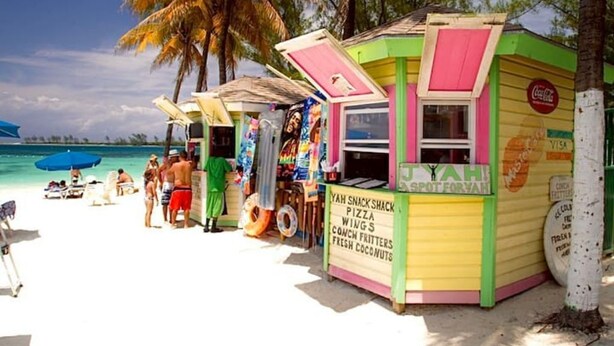 A colorful snack shack on a beach with a sign advertising pizza, wings, and coconut nuts.