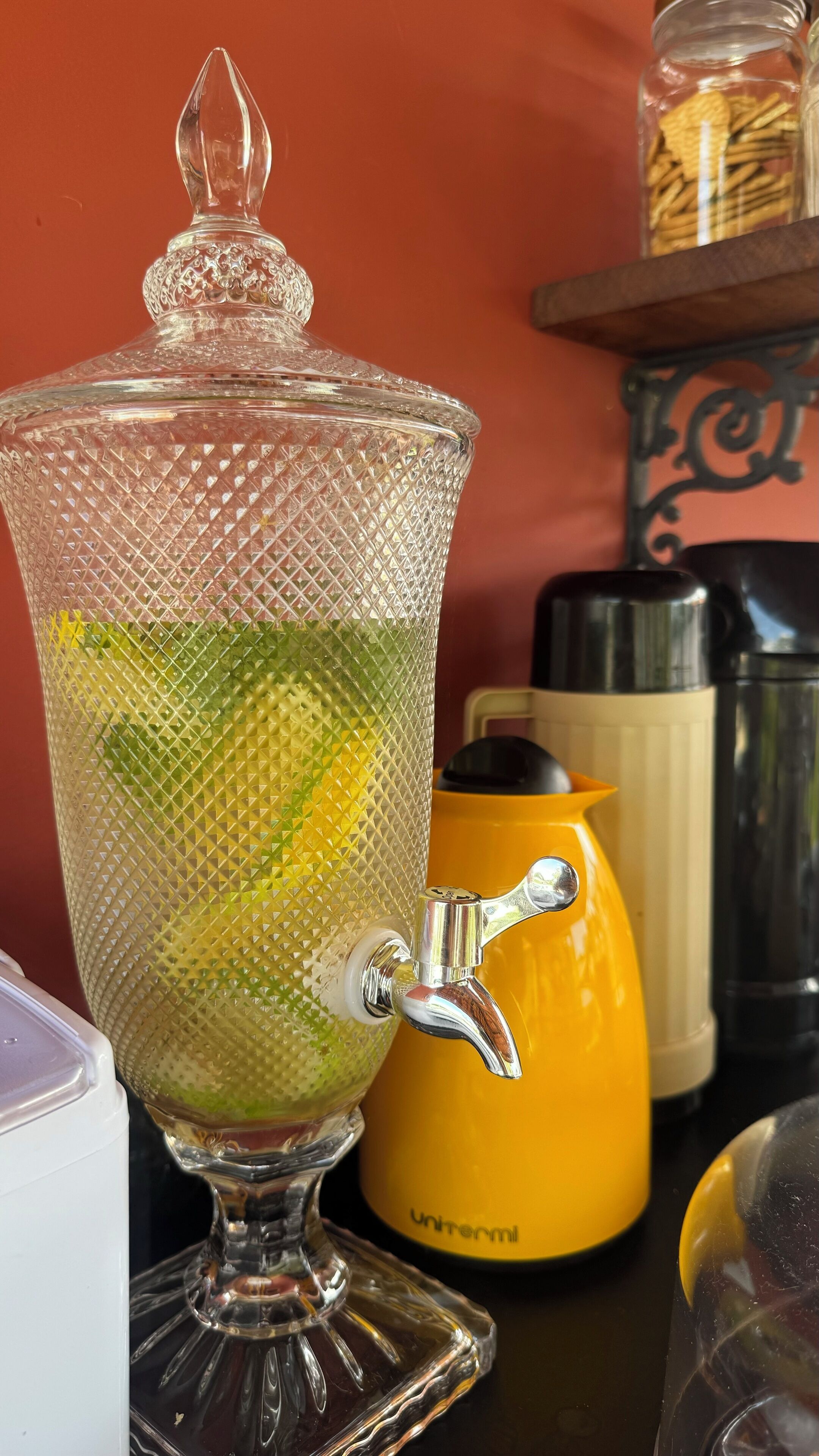 A clear glass teapot with a textured pattern, a yellow water dispenser, and a metal container in the background.