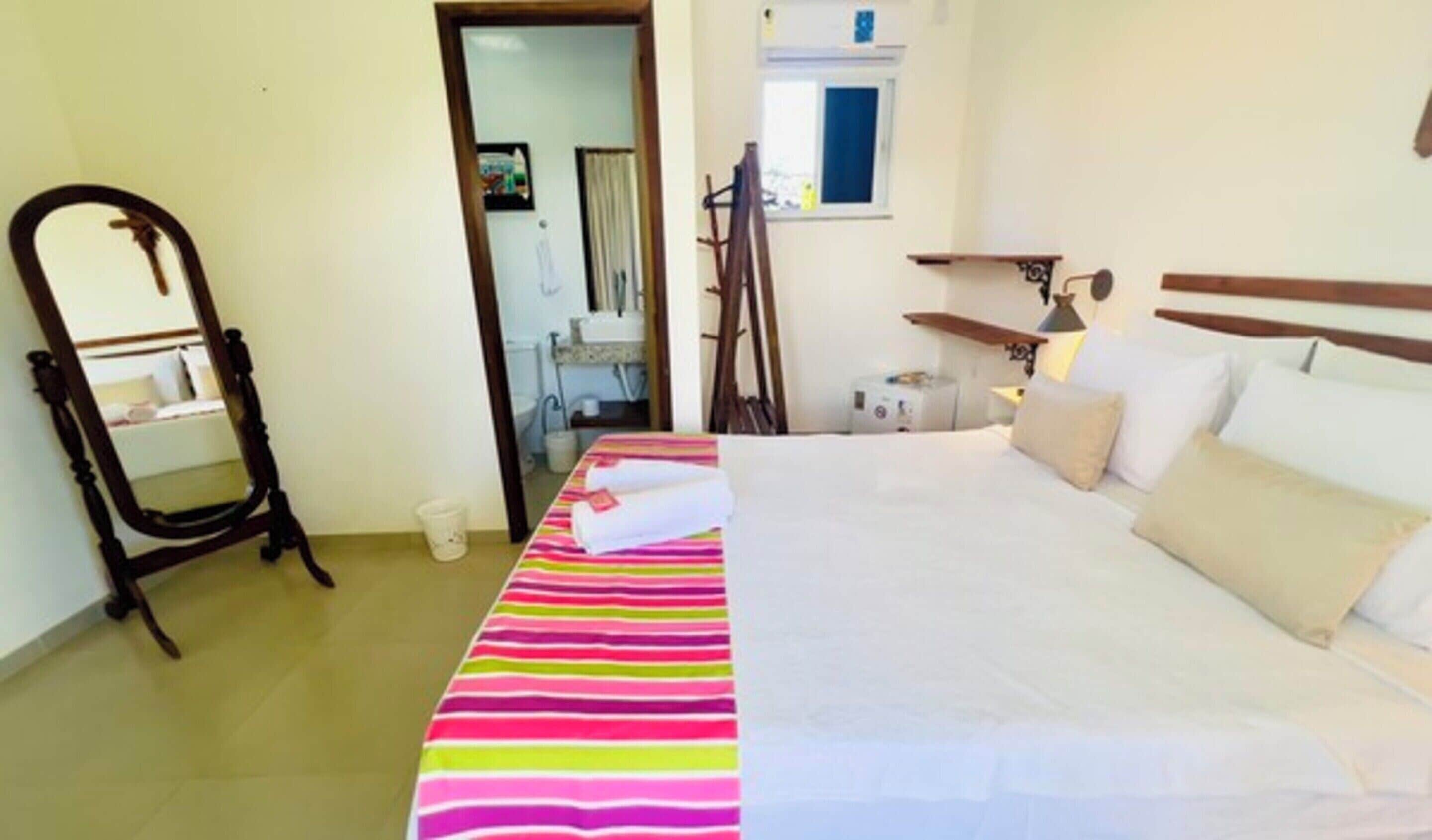 A hotel room with a bed, a mirror, a window, and a small table with a lamp.