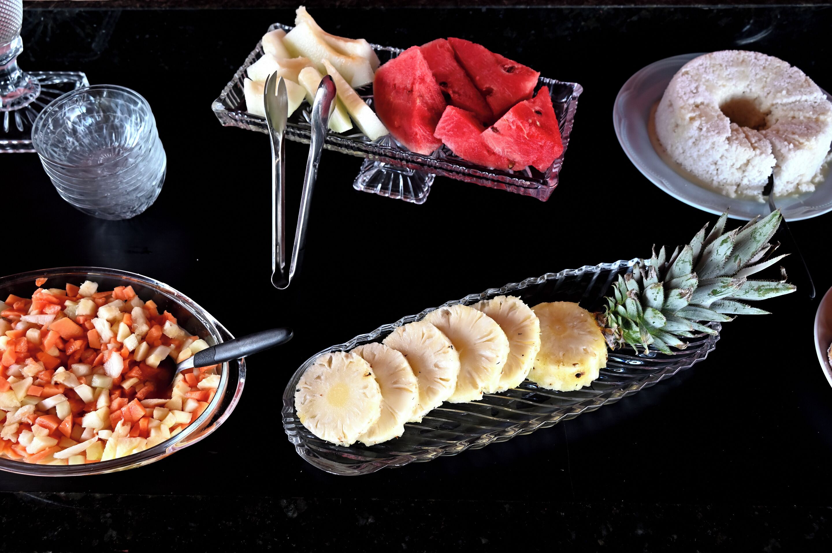 Ein Tisch mit verschiedenen Speisen, darunter eine Schüssel gewürfelten Gemüses, Ananas, Wassermelone und ein Kuchen.