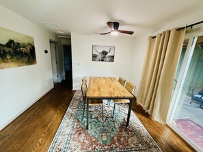 Un comedor con una mesa y sillas de madera, un tapete estampado, un ventilador de techo y un ventanal con cortinas.