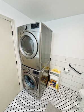 A laundry room with stacked washing machines, a white toilet, and a small shelf with cleaning supplies.
