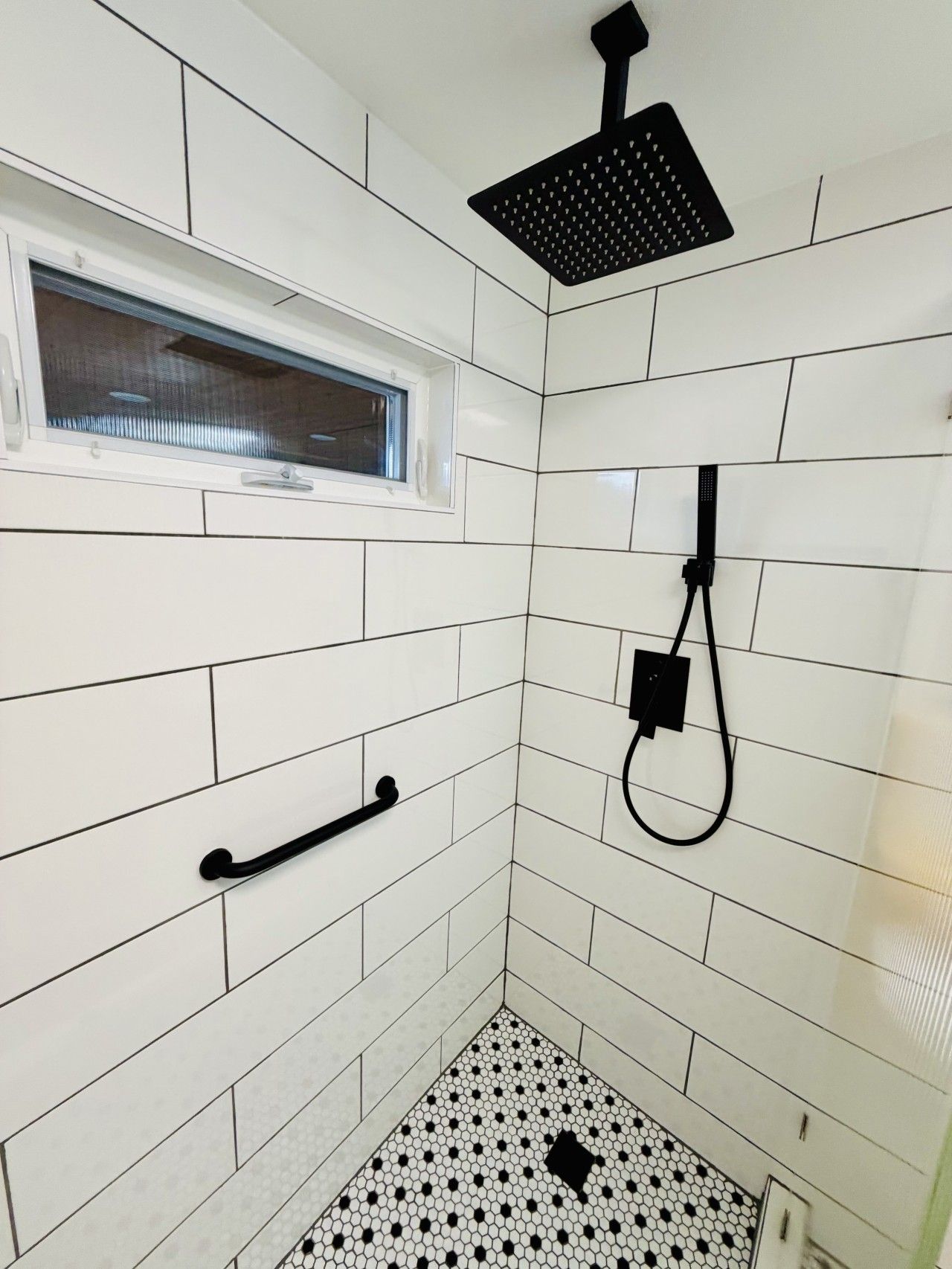 Une petite salle de douche carrelée, dotée d’une fenêtre, d’un pommeau de douche à main et d’un porte-serviettes.