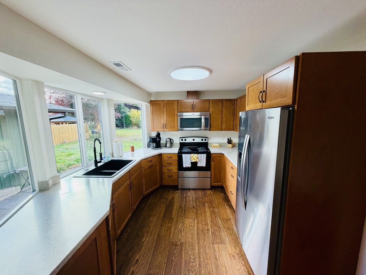 A modern kitchen with wooden cabinets, stainless steel appliances, and a large window providing a view of the outdoors.