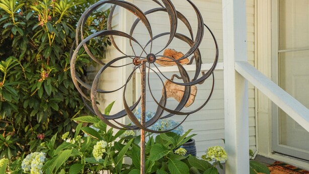 Un carillon éolien en métal, au design rustique, est suspendu à un poteau blanc devant une maison. On aperçoit une fenêtre et une porte à l’arrière-plan.
