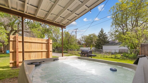 A hot tub under a covered patio with a wooden fence and a grill in the background.