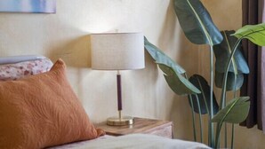 A neatly made bed with a floral bedspread, a single brown pillow, a bedside lamp, and a large green plant.