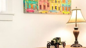 A bedroom with a bed, a nightstand with a lamp, a dresser, and a colorful painting of a street scene. - Bo’s Old West Abode with Hot Tub and Sauna (Billings)