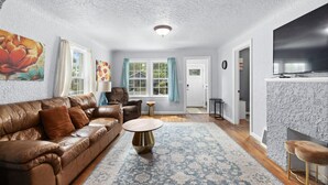 A living room with a brown leather sofa, a round coffee table, and a patterned rug. There is a door leading to another room and a television mounted on the wall.
