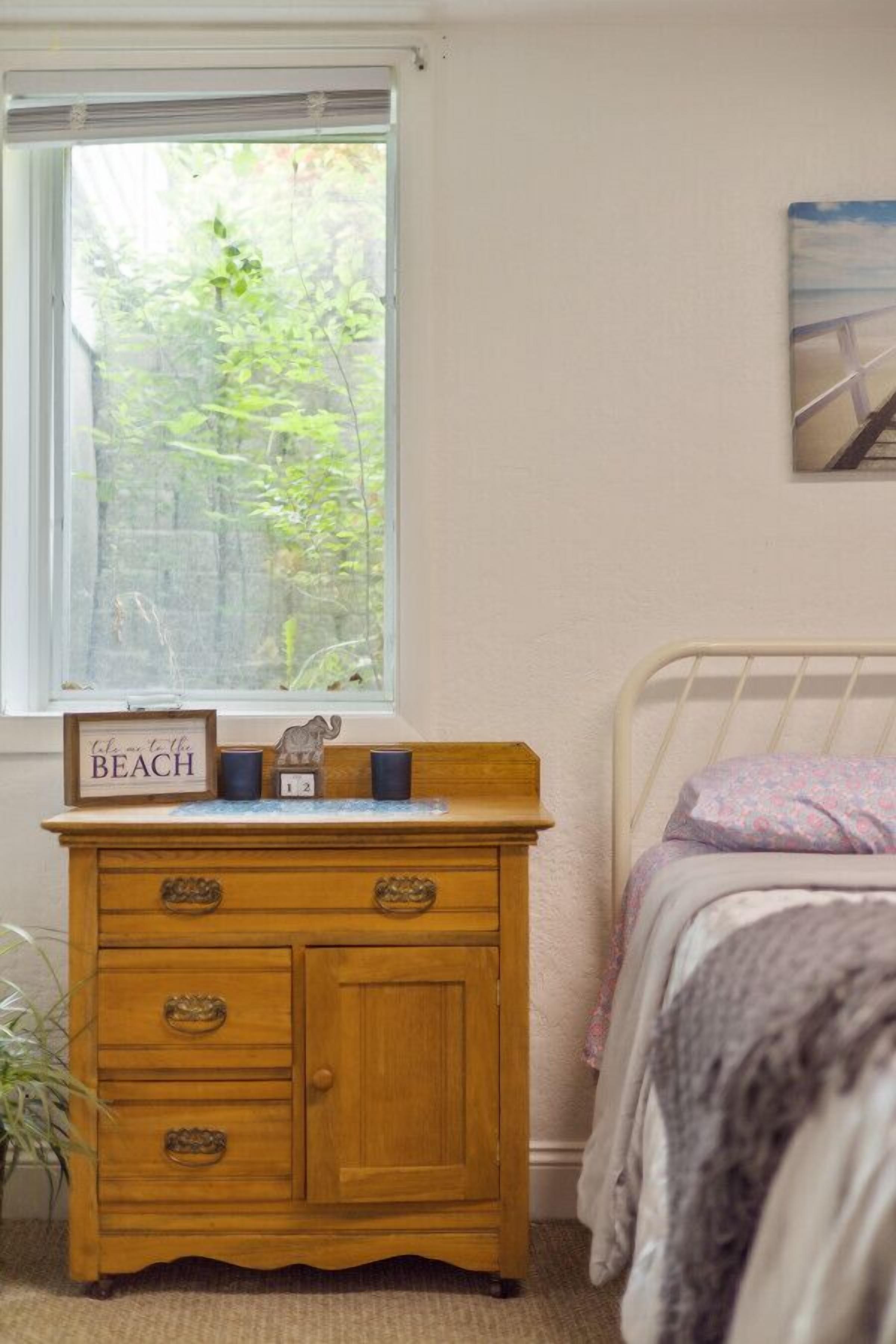 A bedroom with a wooden dresser, a bed with a floral blanket, a window with a view of greenery, and a painting on the wall.