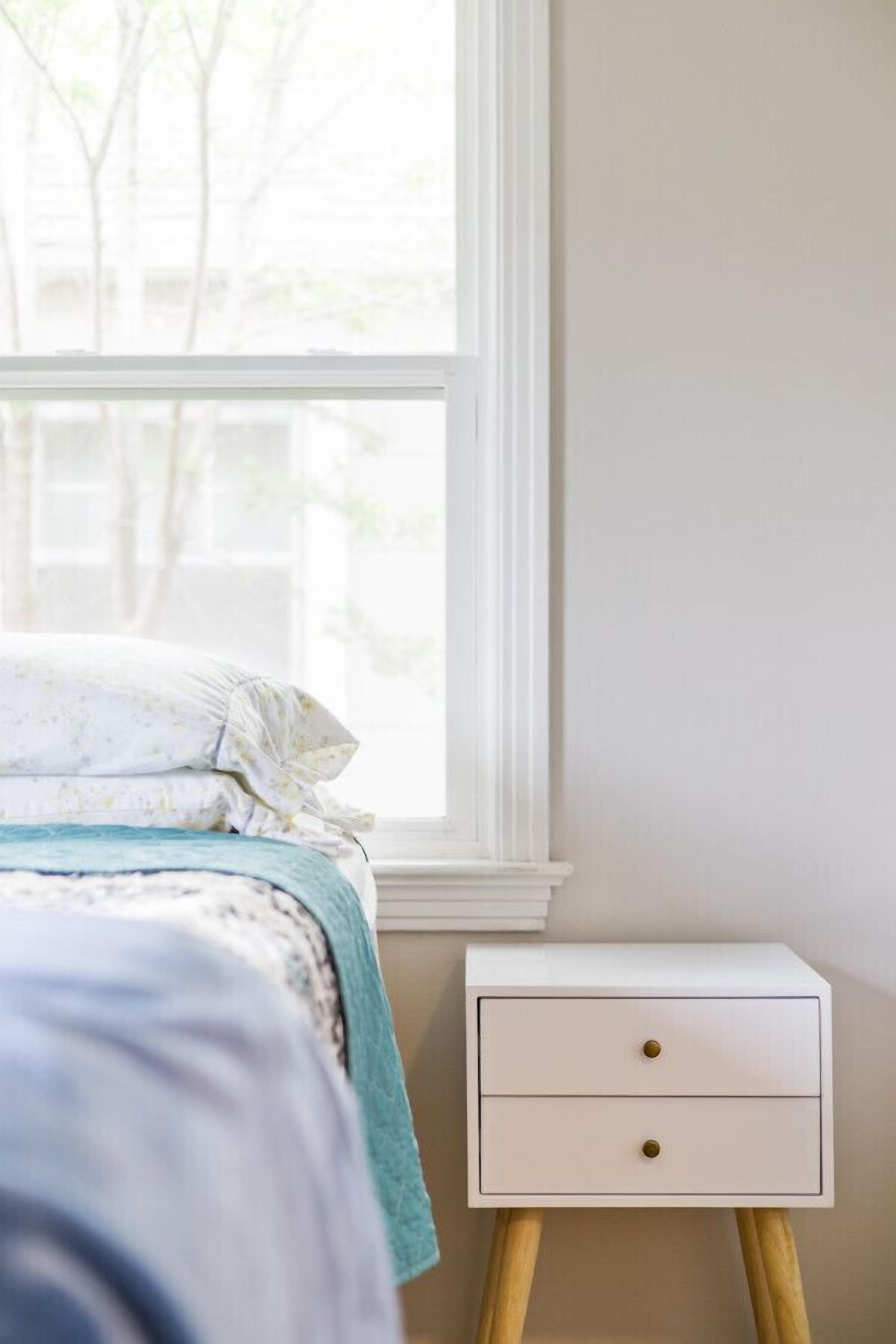 Un lit recouvert d’une couvre-lit à motifs bleus et blancs, une table de chevet blanche avec deux tiroirs et une fenêtre donnant sur des arbres.
