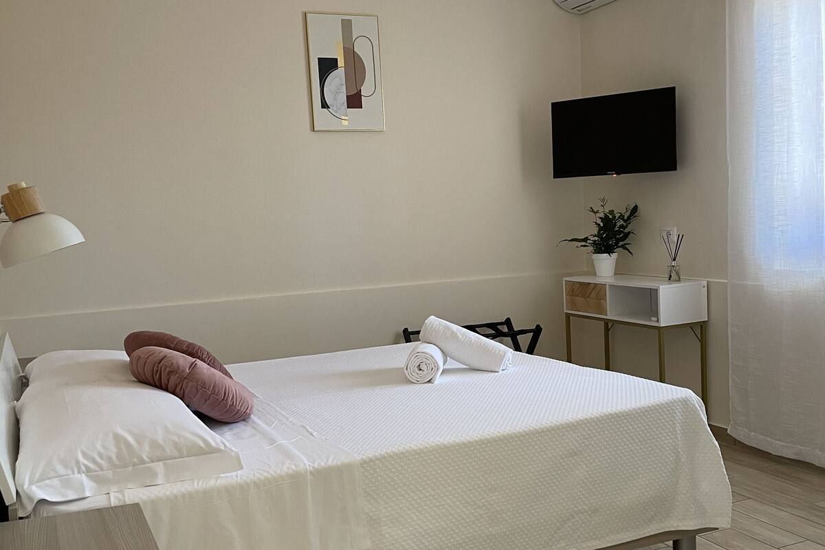 En pænt redt seng med hvide dyne og håndklæde, et lille natbord med en plante, et vægmonteret fjernsyn og en aircondition.