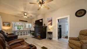 A spacious living room with a fireplace, leather recliners, a ceiling fan, and a kitchen area in the background. - Historic Manor, sleeps 10+, 5 Bedroom, huge yard, lake, fishing, parks, events
 (Winter Haven)
