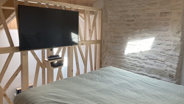 A room with a bed, a mounted TV, and a stone wall. - Le Refuge des Bateliers (Arçais)