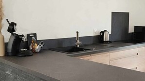 A modern kitchen with a dark countertop, a built-in sink, and a kettle. - LE Mazot-spa-piscine-proche Lac-charme-détente (Lathuile)