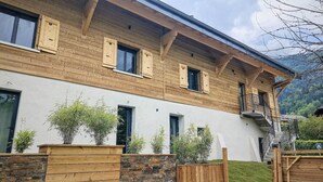 A two-story wooden house with multiple windows, a balcony, and a stone wall in the foreground. - LE Mazot-spa-piscine-proche Lac-charme-détente (Lathuile)