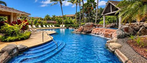 A swimming pool with a waterfall feature, surrounded by palm trees and lounge chairs.