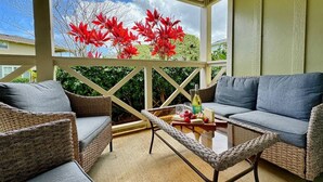 A covered outdoor seating area with wicker furniture and a glass table, overlooking a garden with red foliage.