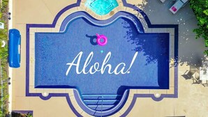 Eine Vogelperspektive auf einen Swimmingpool mit einem 'Aloha!'-Schild, umgeben von einer Fliesenveranda und einem kleinen Whirlpool.