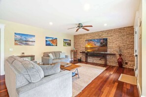 Ein Wohnzimmer mit grauem Sofa, einem Holztisch, einem Fernseher an einer Steinwand und einem Deckenventilator.