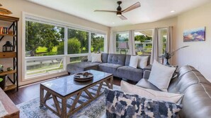 A spacious living room with a large window, a ceiling fan, and a sofa with cushions. - Deluxe Princeville Condo! Golf Views & Heated Pool (Princeville)