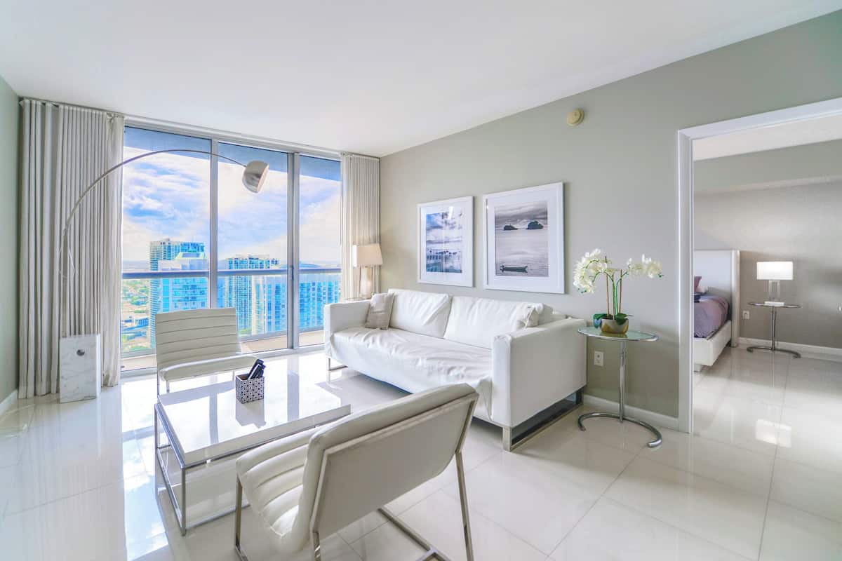 Quarto de hotel moderno com varanda, um sofá branco, uma mesa de jantar com cadeiras e uma pequena mesa lateral com um vaso de flores.