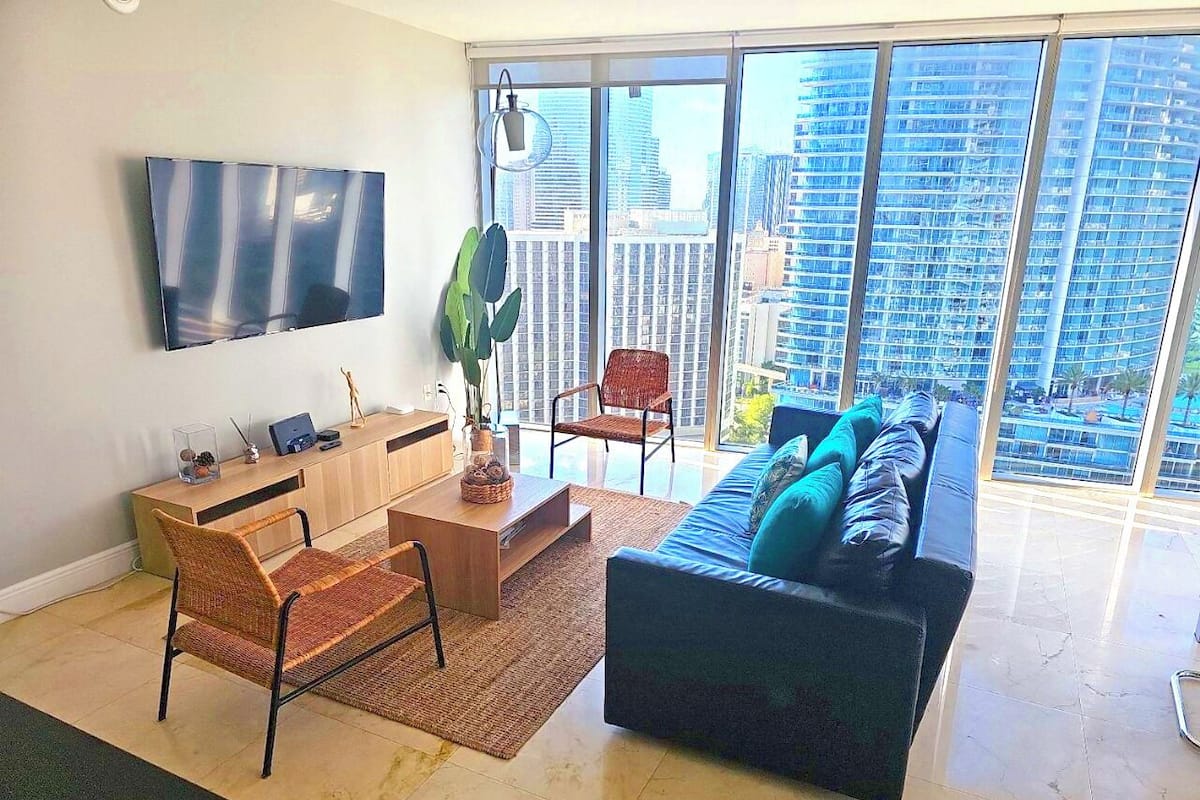 Uma sala de estar moderna com uma janela ampla, uma televisão de tela plana, um sofá, uma mesa de centro e uma escrivaninha com um computador.