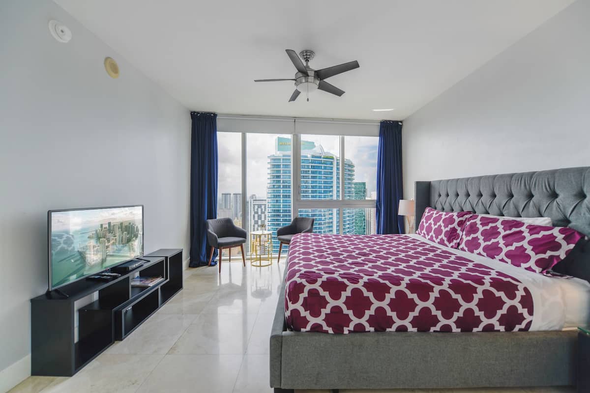 Quarto de hotel moderno com uma cama grande, televisão, ventoinha no teto e vista para o horizonte urbano através de grandes janelas.