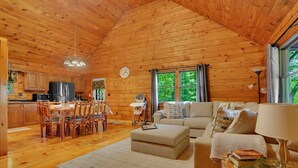 A wooden cabin interior with a dining area, kitchen, living room, and a large window with curtains. - Unplug from the daily grind, whether a fun family getaway or a retreat with friends, this cabin is a perfect refuge to unwind and rejuvenate. Brand new Hot Tub! (Elliottsburg)