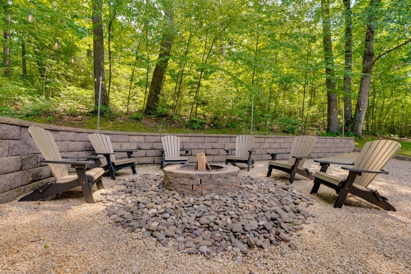 Un espace de détente circulaire, avec un foyer au centre, entouré de fauteuils Adirondack, le tout adossé à un mur de pierres et surplombant une forêt dense.
