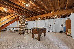 A spacious room with a wooden ceiling, a brick pillar, a foosball table, a table with chairs, and a staircase.