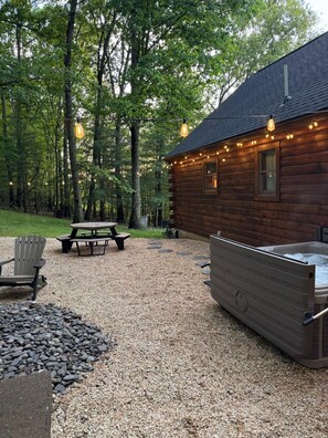 A log cabin with a hot tub, picnic table, and outdoor seating area surrounded by a gravel path and trees.