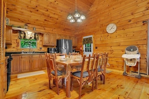 A wooden cabin interior with a dining table, chairs, a refrigerator, a microwave, and a clock on the wall.