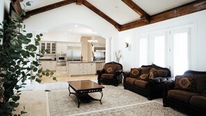 A spacious living room with a high ceiling, wooden beams, a fireplace, and a view into the kitchen.