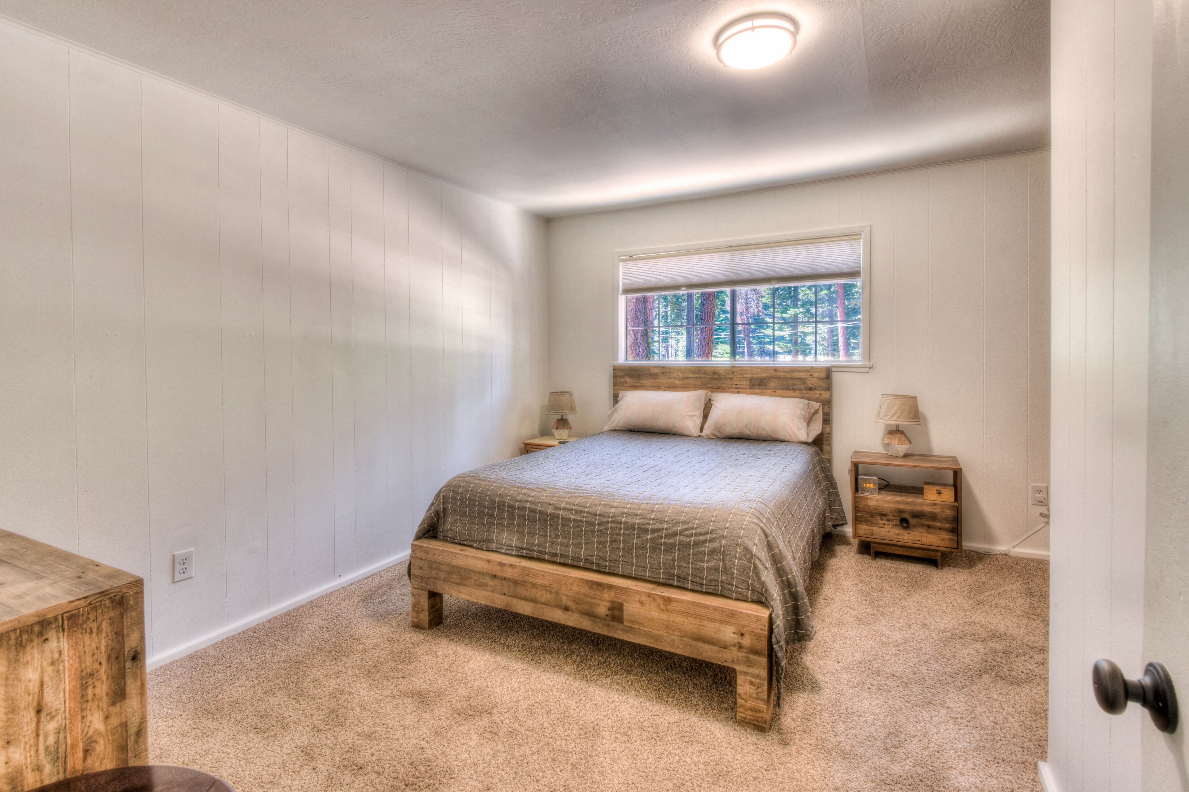 A bedroom with a wooden bed, a nightstand, a window with a view of trees, and a door.