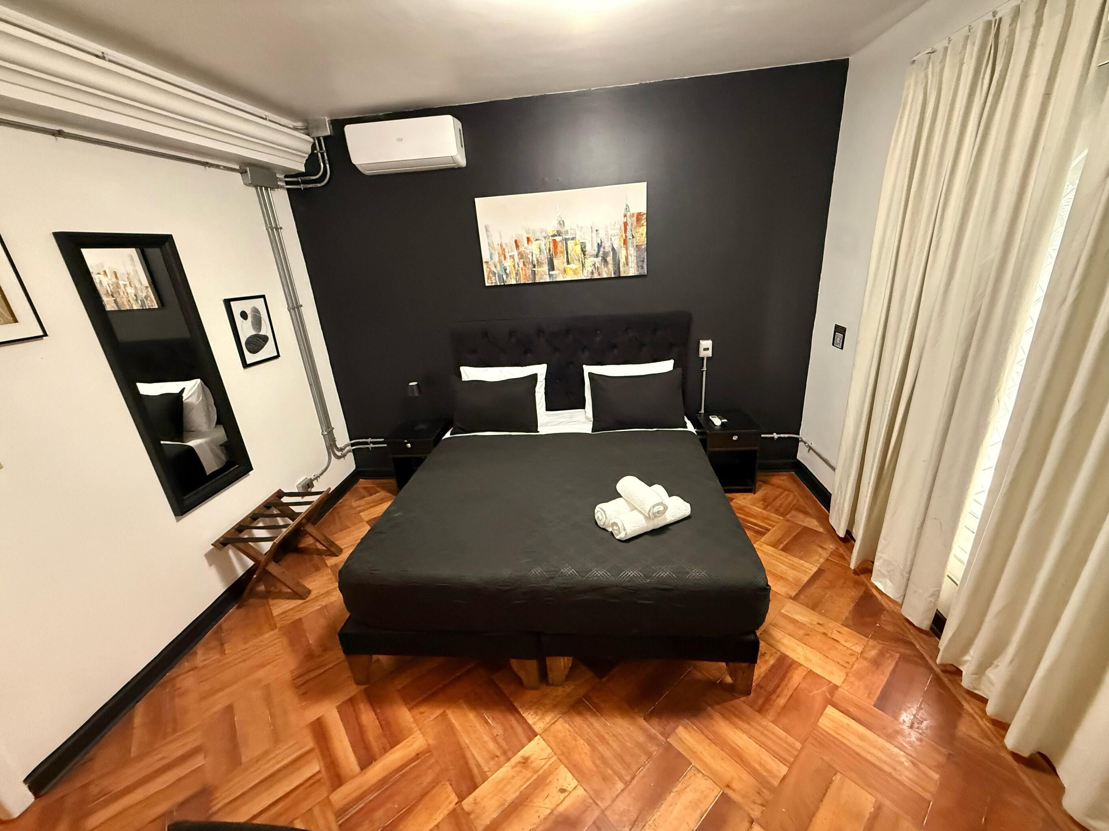 A hotel room with a large bed, a painting on the wall, a mirror, and a window with curtains.