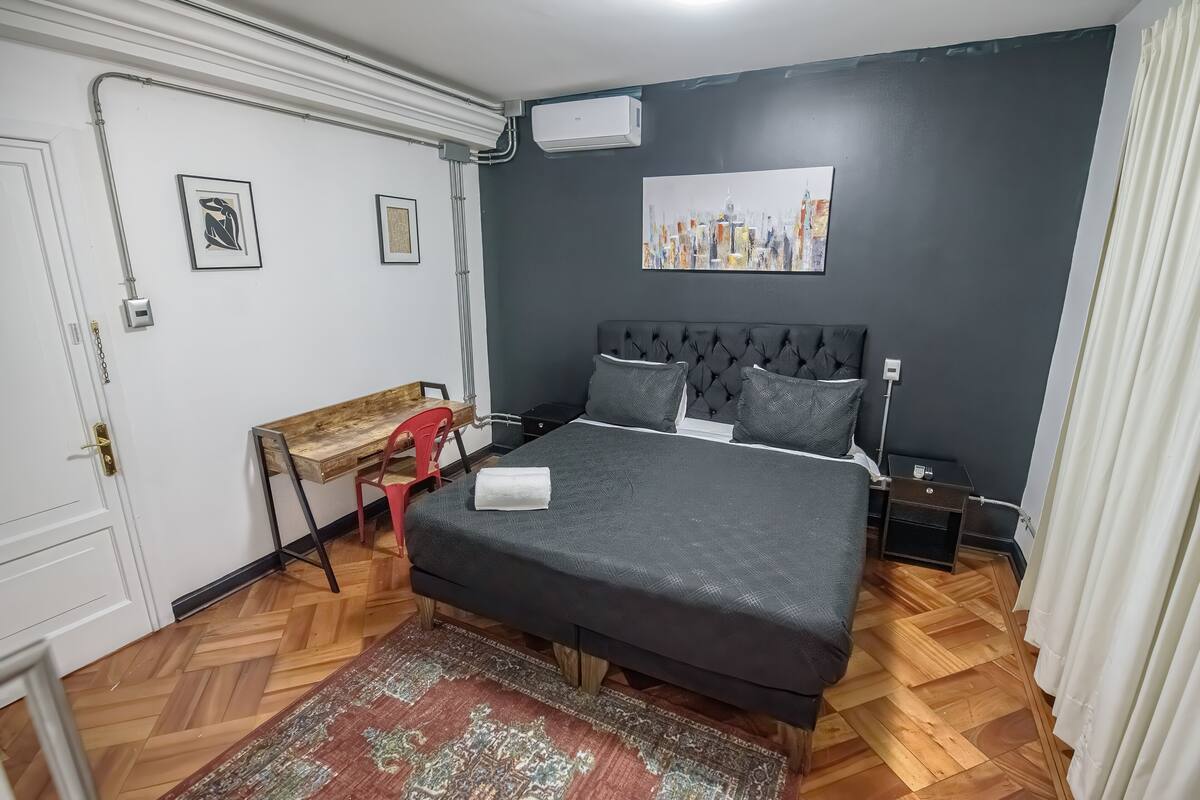 A bedroom with a bed, a desk, a chair, a nightstand, and a wall-mounted air conditioner.