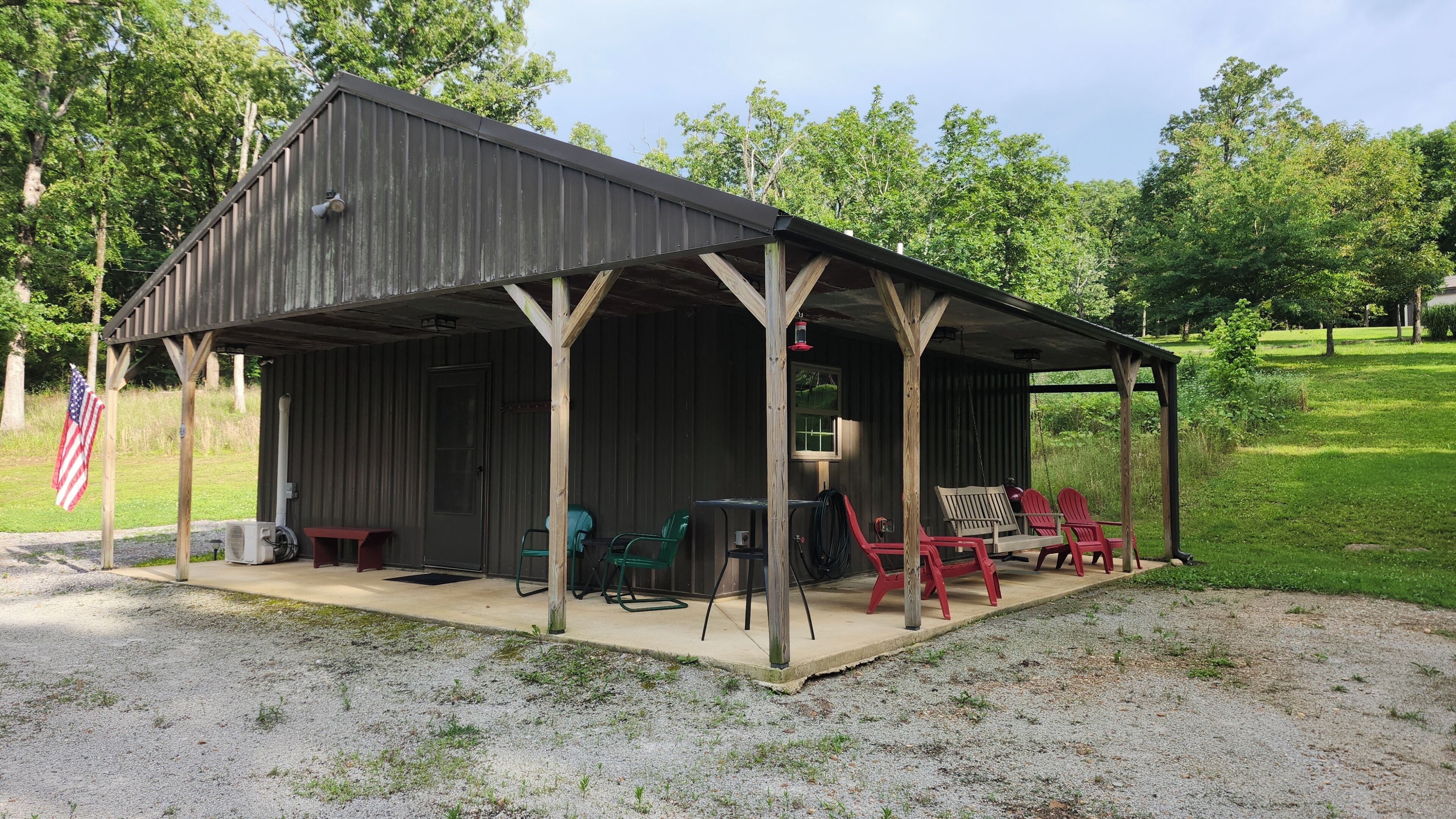 A covered outdoor seating area with chairs and a table, an American flag, and a building with a corrugated metal roof.