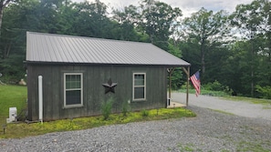 A small wooden cabin with a metal roof, two windows with shutters, a star decoration, and an American flag.