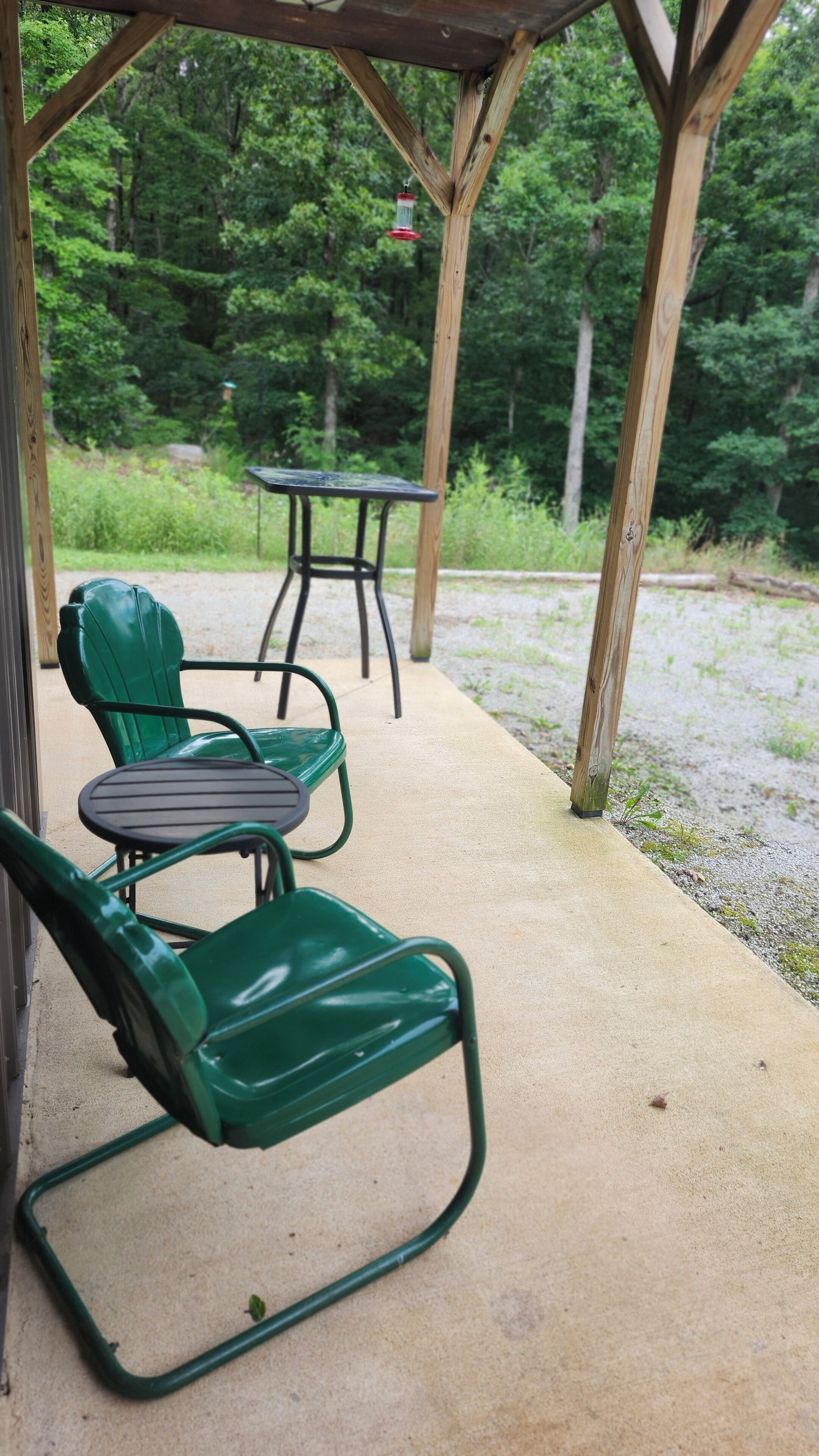 A covered patio area with two green chairs and a table, surrounded by a wooden pergola and a gravel path.