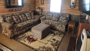 Ein gemütliches Blockhäuschen-Interieur mit einem floralen Sofa, einem kleinen Hocker, einem Holztisch, einer Uhr und einem Deckenventilator.
