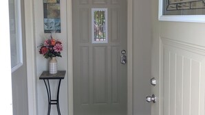 A front door with a 'HELLO' mat, a small table with a vase of flowers, and a wall-mounted light fixture. - Sweet One Bedroom in the heart of the Keweenaw! (Ahmeek)