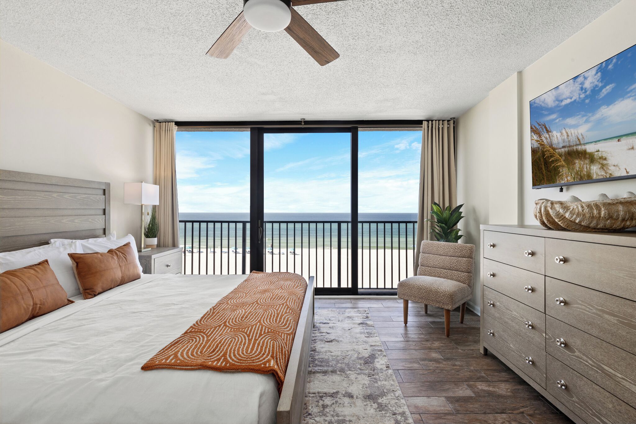 Une chambre d’hôtel avec un grand lit, un ventilateur de plafond, une vue sur la plage, une chaise et une commode en bois.