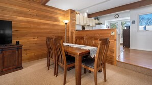 A wooden dining table with chairs, a television on a cabinet, and a kitchen area in the background. - Villages of Wisp 14 Winding Way by Taylor Made Deep Creek (McHenry)