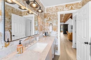Features a spacious bathroom with a large mirror, sink, and countertop, surrounded by patterned wallpaper and a door to an adjacent room.