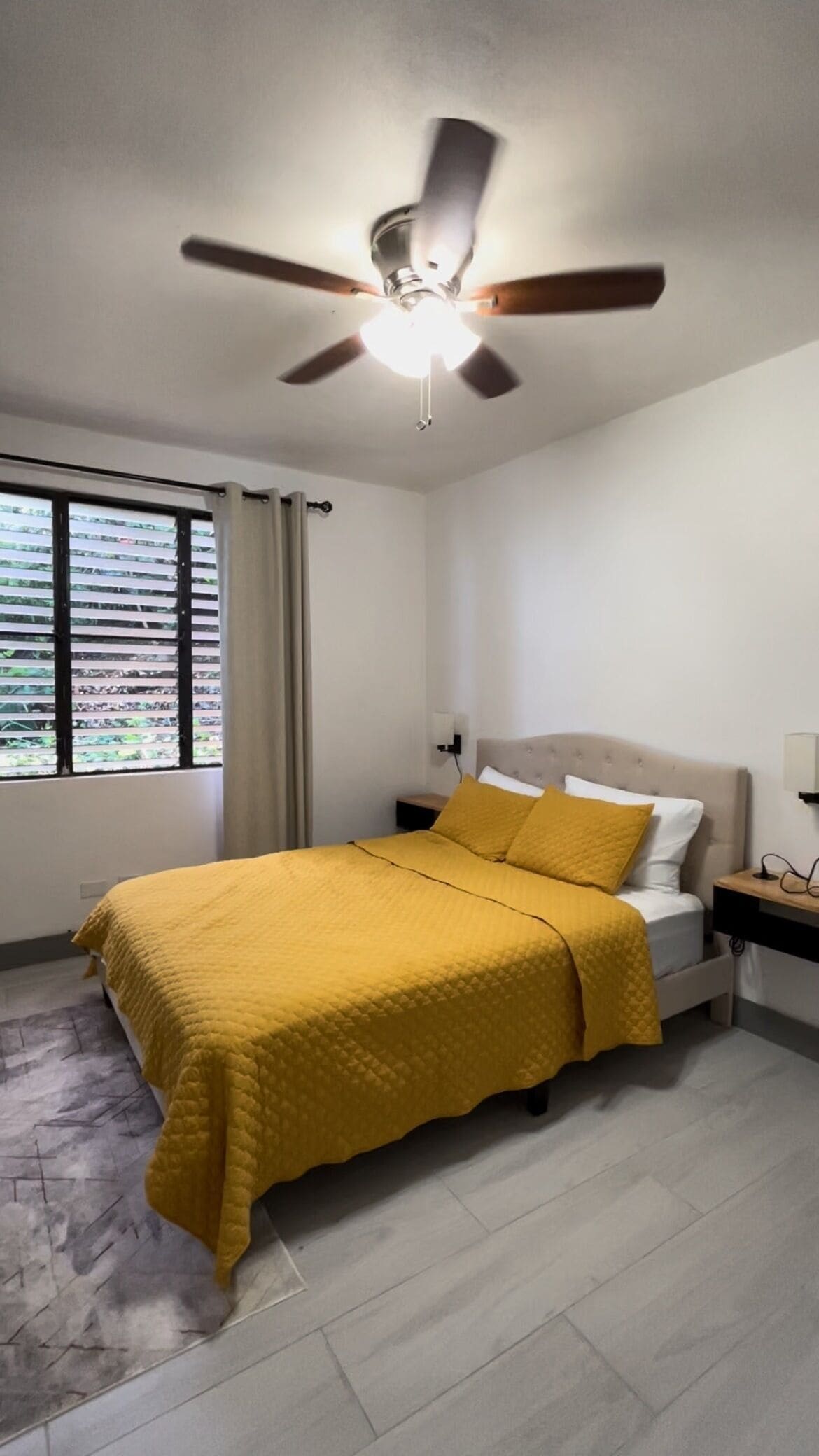 A bedroom with a bed, a ceiling fan, a nightstand, and a window with blinds.
